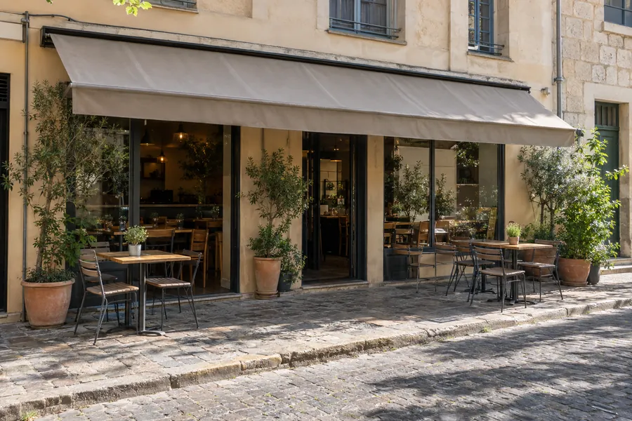 A small restaurant with a terrace and a plain awning, seen from across a quiet street on a sunny morning