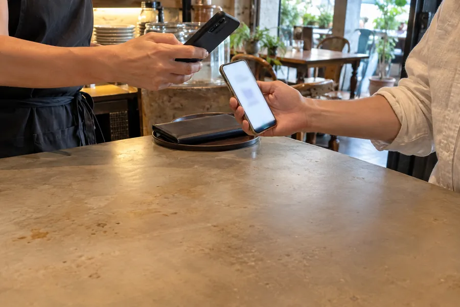 At a restaurant counter, a staff member's phone scanning the phone a guest holds out