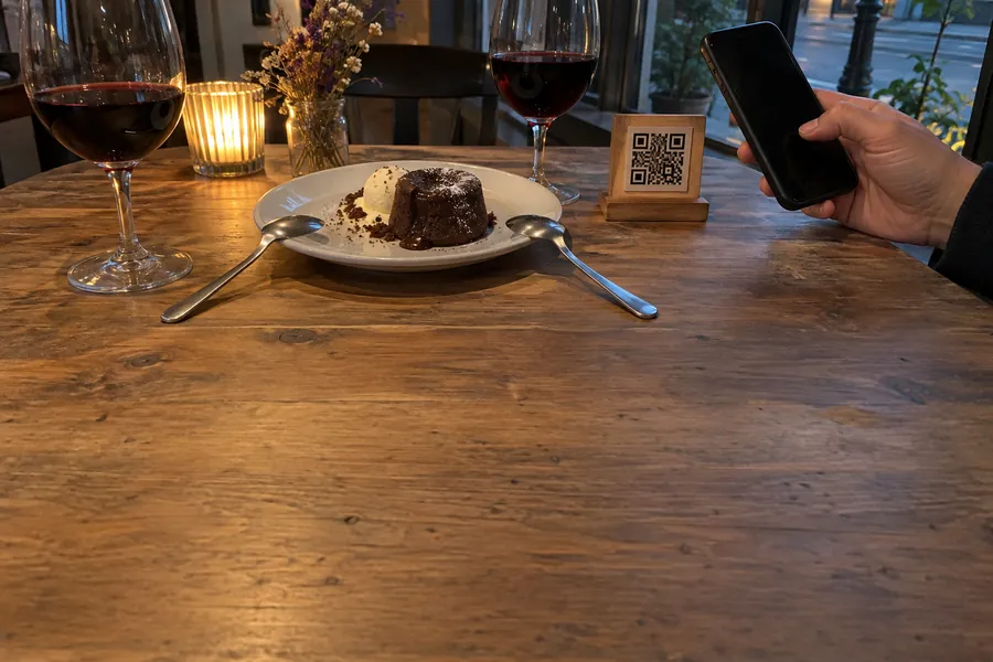 A chocolate fondant, two glasses of wine and a small QR code stand on a bistro table, a hand holding a phone