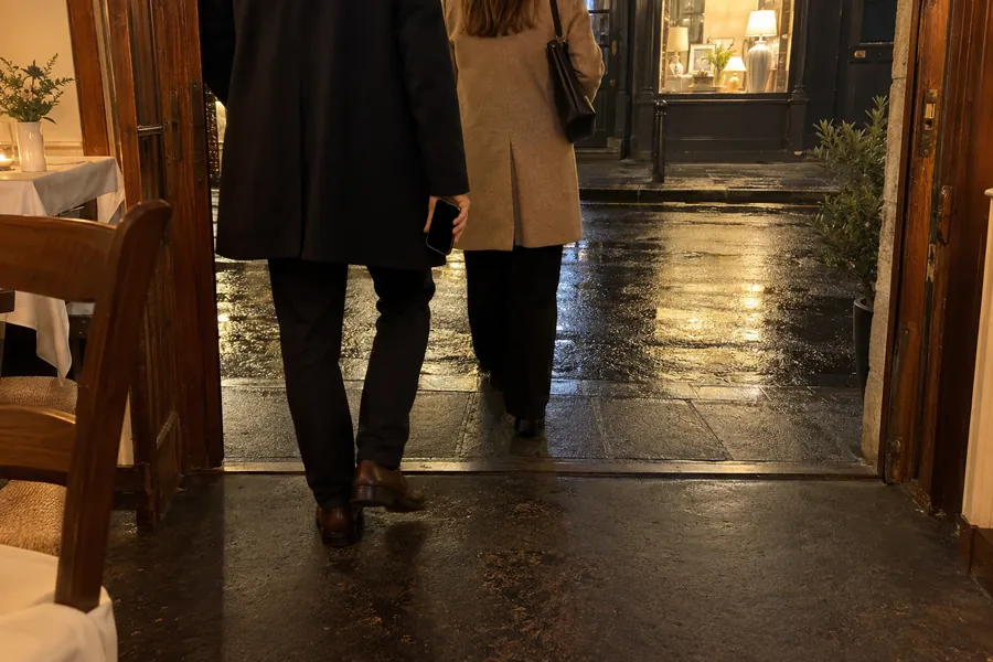 Guests in coats leaving a small bistro late in the evening, seen from behind, one holding a phone, warm light from the dining room
