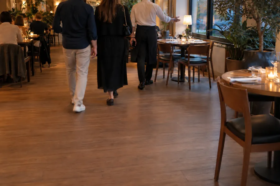 A waiter leading two arriving guests between the tables of a busy bistro in the evening