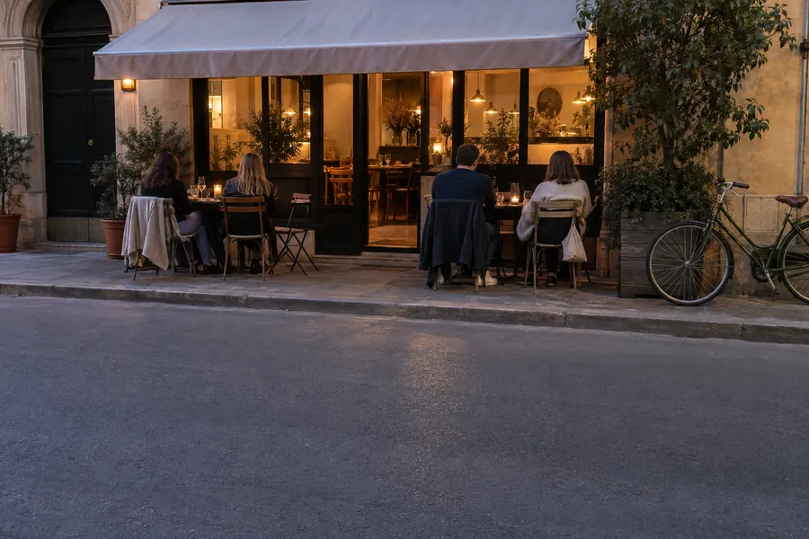 A small restaurant at dusk seen from across the street, warm light in the windows and guests on the terrace