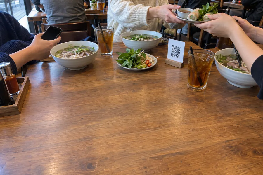 Friends at lunch in a Vietnamese restaurant, one holding a phone over bowls of pho next to a small QR code stand