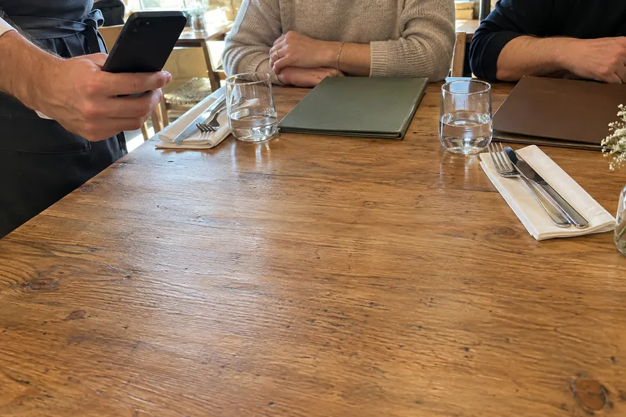 A waiter holding a phone at the edge of a lunch table, guests' hands resting next to glasses and menus